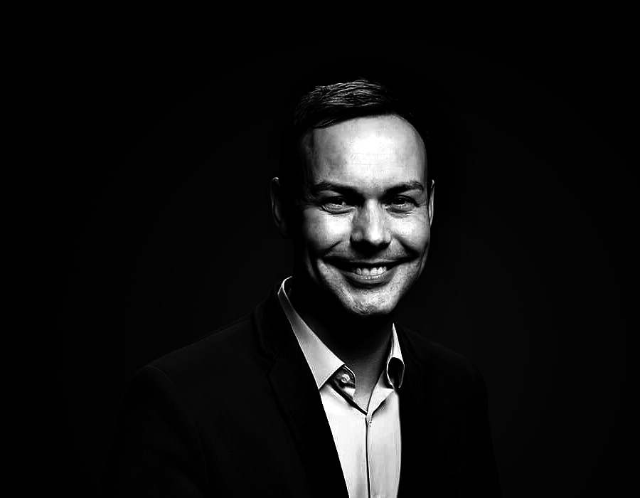 Francis Hellyer, smiling, in a dark jacket and open-collared shirt, photographed in black and white against a black background